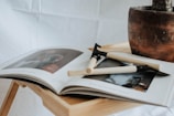 An open magazine lies on a wooden surface, displaying images in muted tones. Two beige-handled safety razors rest diagonally across the pages. A rustic, brown ceramic pot sits on the table, partially in view.