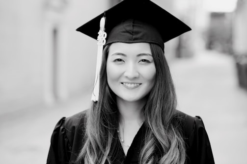 A woman in a graduation cap and gown standing outdoors on a blurred street background. She has long hair and is smiling at the camera.