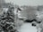 A snowy suburban landscape featuring several houses with white rooftops blanketed by snow. Tall, evergreen trees and bare deciduous trees are covered in snow, as well as a garden area with a swing set. The ground is entirely covered in white snow, creating a serene winter scene.