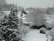 A snowy suburban landscape featuring several houses with white rooftops blanketed by snow. Tall, evergreen trees and bare deciduous trees are covered in snow, as well as a garden area with a swing set. The ground is entirely covered in white snow, creating a serene winter scene.