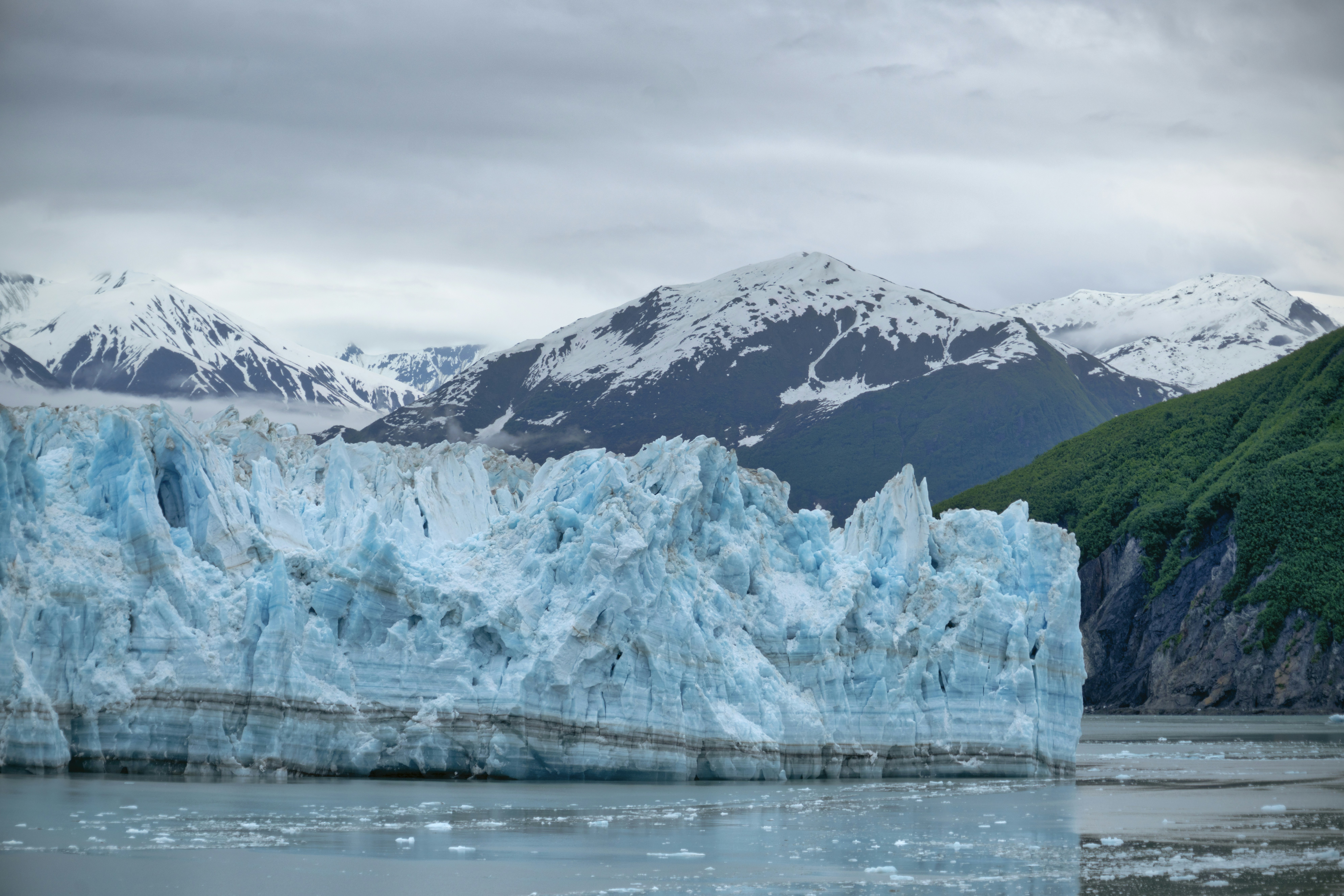 A photo of Hubbard Glacier in Alaska, featuring a massive tidewater glacier with a brown and white surface ending at a calm blue body of water. The glacier is surrounded by snow-capped mountains in the distance. (Photo by Peter Hansen on Unsplash)