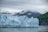 A vast glacier stretches across the foreground, with its jagged ice formations and bluish hues contrasting against the surrounding snow-covered mountains. The sky is overcast, casting a muted light over the scene. To the right, lush green hills border the glacier, adding a touch of vivid color to the otherwise cool-toned landscape.