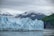 A vast glacier stretches across the foreground, with its jagged ice formations and bluish hues contrasting against the surrounding snow-covered mountains. The sky is overcast, casting a muted light over the scene. To the right, lush green hills border the glacier, adding a touch of vivid color to the otherwise cool-toned landscape.