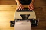 A close-up of hands typing on a vintage typewriter with scattered papers around.