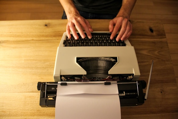 A pair of hands typing on a white typewriter placed on a wooden table. The typewriter has a sheet of paper inserted, ready for typing. The setting suggests a retro or vintage feel.