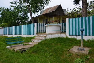 A small, rustic house with a metal roof and a green and white picket fence surrounding it. In front of the house is a concrete well with a metal railing around it. A green wooden bench is positioned on a concrete slab in the grassy yard. A pipe structure, possibly part of the well system, is visible to the right. Trees and shrubs add greenery, creating a peaceful, rural setting.