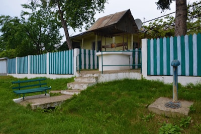 A small, rustic house with a metal roof and a green and white picket fence surrounding it. In front of the house is a concrete well with a metal railing around it. A green wooden bench is positioned on a concrete slab in the grassy yard. A pipe structure, possibly part of the well system, is visible to the right. Trees and shrubs add greenery, creating a peaceful, rural setting.
