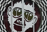 A large graphic of a stylized face with prominent oversized glasses displaying the word 'GO'. The image is part of a glass door with a surrounding black and white checkered circular pattern. The facial features are minimalist and cartoon-like, presenting a playful and eye-catching design.