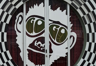 A large graphic of a stylized face with prominent oversized glasses displaying the word 'GO'. The image is part of a glass door with a surrounding black and white checkered circular pattern. The facial features are minimalist and cartoon-like, presenting a playful and eye-catching design.