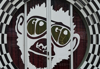 A large graphic of a stylized face with prominent oversized glasses displaying the word 'GO'. The image is part of a glass door with a surrounding black and white checkered circular pattern. The facial features are minimalist and cartoon-like, presenting a playful and eye-catching design.