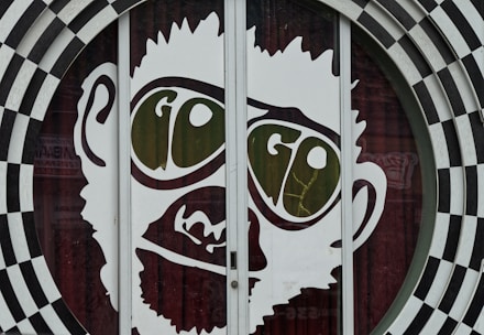 A large graphic of a stylized face with prominent oversized glasses displaying the word 'GO'. The image is part of a glass door with a surrounding black and white checkered circular pattern. The facial features are minimalist and cartoon-like, presenting a playful and eye-catching design.