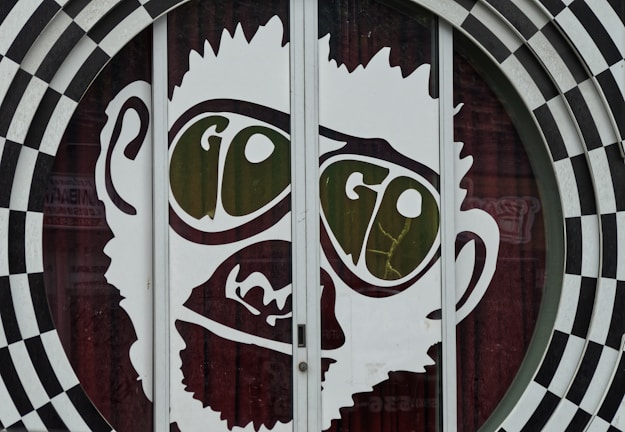 A large graphic of a stylized face with prominent oversized glasses displaying the word 'GO'. The image is part of a glass door with a surrounding black and white checkered circular pattern. The facial features are minimalist and cartoon-like, presenting a playful and eye-catching design.