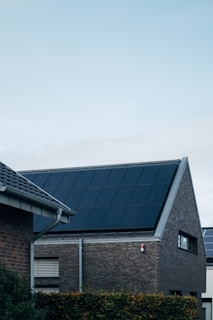 A modern triangular-roofed building with large solar panels is situated against a light blue sky. The structure appears to be made of dark brick with additional small windows, alongside other nearby buildings. A green hedge lines the garden area in front of the house.