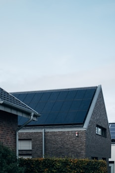 A modern triangular-roofed building with large solar panels is situated against a light blue sky. The structure appears to be made of dark brick with additional small windows, alongside other nearby buildings. A green hedge lines the garden area in front of the house.