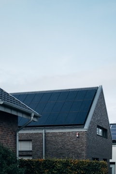A modern triangular-roofed building with large solar panels is situated against a light blue sky. The structure appears to be made of dark brick with additional small windows, alongside other nearby buildings. A green hedge lines the garden area in front of the house.