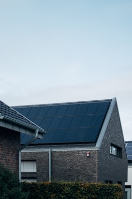 A modern triangular-roofed building with large solar panels is situated against a light blue sky. The structure appears to be made of dark brick with additional small windows, alongside other nearby buildings. A green hedge lines the garden area in front of the house.