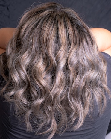 Wavy, multi-toned hair flowing down the back of a person. The hair features shades of brown and blonde, creating a natural and voluminous look.