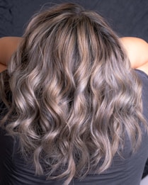Wavy, multi-toned hair flowing down the back of a person. The hair features shades of brown and blonde, creating a natural and voluminous look.