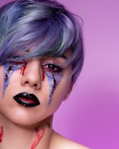 A person with short, multicolored hair, predominantly purple and blue, has dramatic makeup. The makeup includes bold, dark lipstick, glittery eyeshadow with streaks of red resembling tears, and red markings on the neck. The background is a solid light purple.