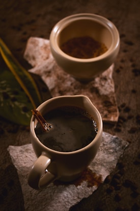 Two ceramic cups are placed on textured surfaces, with one filled with dark coffee and a cinnamon stick. Scattered coffee beans and green leaves create a rustic ambiance.