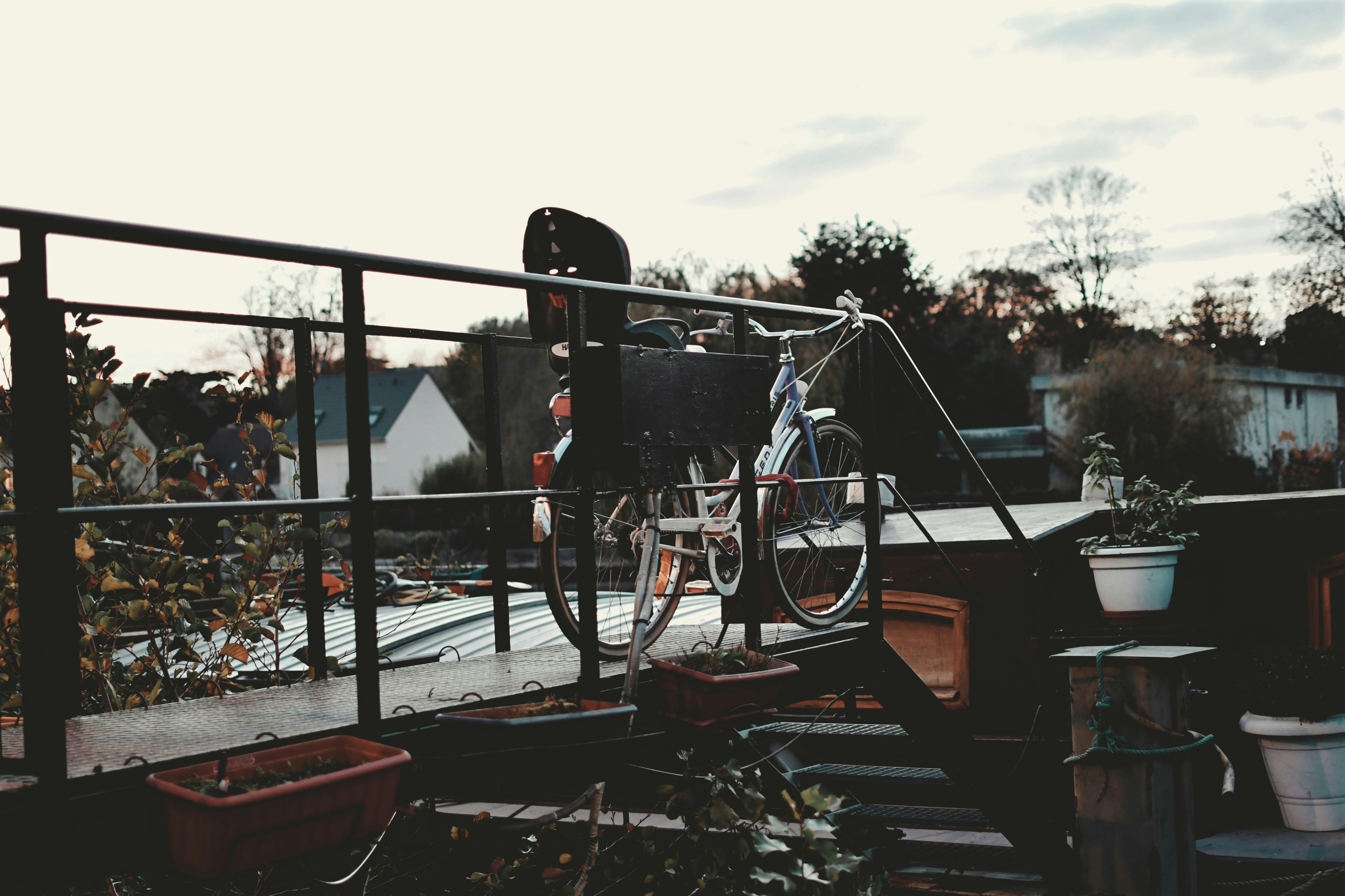 A bicycle, child seat, and potted plants on a boat deck, framed by dark railings, with distant houses and trees under a muted, tranquil sky.