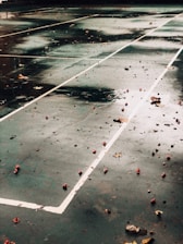 A wet, empty tennis court with scattered leaves and debris on the surface. White boundary lines create geometric patterns on the dark green ground. The surface is glossy from recent rain, reflecting darker shades.
