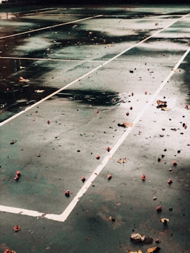 A wet, empty tennis court with scattered leaves and debris on the surface. White boundary lines create geometric patterns on the dark green ground. The surface is glossy from recent rain, reflecting darker shades.