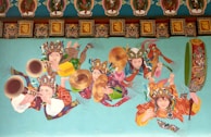 A vibrant and colorful mural depicts a group of people in traditional attire playing musical instruments. The instruments include large windpipes, drums, and cymbals. The individuals are adorned with ornate headpieces and intricate costumes featuring rich patterns and multiple colors. Above them, decorative patterns and artwork add to the elaborate scene.