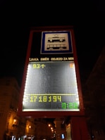 A digital street sign displays public transportation information with tram and bus symbols. The sign includes scrolling text in Czech, with LED numbers and letters in yellow and green against a dark urban background. Streetlights illuminate the nearby buildings and trees.