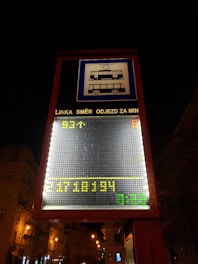 A digital street sign displays public transportation information with tram and bus symbols. The sign includes scrolling text in Czech, with LED numbers and letters in yellow and green against a dark urban background. Streetlights illuminate the nearby buildings and trees.