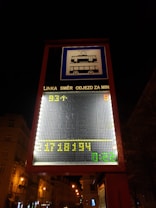 A digital street sign displays public transportation information with tram and bus symbols. The sign includes scrolling text in Czech, with LED numbers and letters in yellow and green against a dark urban background. Streetlights illuminate the nearby buildings and trees.