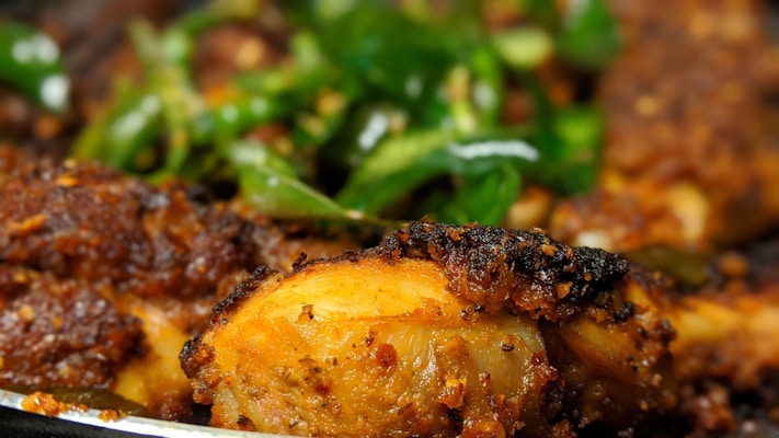 A close-up of a piece of grilled or roasted chicken with a crispy, seasoned exterior. The chicken appears to be garnished with fresh green herbs, possibly cilantro or green onions, providing a contrast of colors and textures.