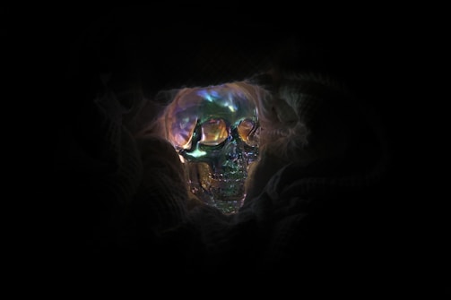 A luminous, multicolored skull appears amidst dark shadows. The skull's surface is reflective with iridescent hues, including shades of blue, green, and orange. It is surrounded by smoky, ethereal textures, enhancing the mysterious atmosphere.
