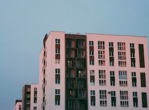 A modern apartment building features a minimalist design with clean lines and large windows. The structure showcases a combination of white and dark brown colors, with each apartment unit having its own balcony or window. The sky is clear, adding a serene backdrop to the urban architecture.