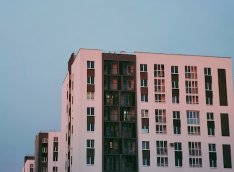 A modern apartment building features a minimalist design with clean lines and large windows. The structure showcases a combination of white and dark brown colors, with each apartment unit having its own balcony or window. The sky is clear, adding a serene backdrop to the urban architecture.