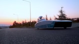 A sleek, futuristic solar-powered car is parked on the side of a road at sunset. The sky is filled with hues of orange and pink, and the surrounding area is lined with trees and streetlights. The vehicle has a streamlined design with a transparent canopy.