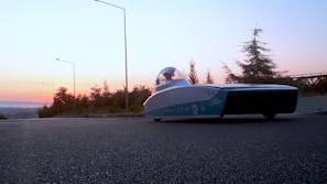 A sleek, futuristic solar-powered car is parked on the side of a road at sunset. The sky is filled with hues of orange and pink, and the surrounding area is lined with trees and streetlights. The vehicle has a streamlined design with a transparent canopy.