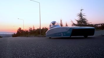 A sleek, futuristic solar-powered car is parked on the side of a road at sunset. The sky is filled with hues of orange and pink, and the surrounding area is lined with trees and streetlights. The vehicle has a streamlined design with a transparent canopy.