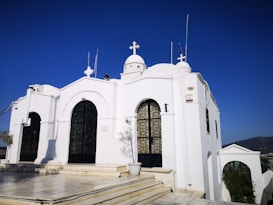 A white building with arched doorways and windows, topped with a cross, stands prominently. The structure has a series of domes and antennas on the roof. A potted plant sits on the tiled walkway leading to the building, under a clear blue sky.