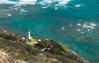 Aerial view of a bright white lighthouse with a red top located on a green peninsula, surrounded by the turquoise waters of the ocean. The coastline is lined with trees and small structures, while waves crash gently onto the shore.
