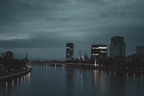 A cityscape during dusk with modern skyscrapers and a river in the foreground. The sky is overcast with dark clouds, and the buildings are illuminated with artificial lights reflecting on the water. A bridge is visible in the distance, connecting the two sides of the river.