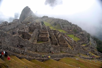 Ancient stone ruins extend across a mountainous landscape, shrouded in mist with terraced greenery visible between the structures. Small groups of people are scattered throughout, exploring the historic site.