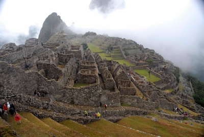Ancient stone ruins extend across a mountainous landscape, shrouded in mist with terraced greenery visible between the structures. Small groups of people are scattered throughout, exploring the historic site.
