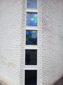 A corner of a brick building with several vertically aligned blue and green stained glass windows. The bricks are white and the windows have abstract designs composed of various shades of blue and green glass pieces.