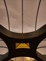 The image features an architectural structure with a series of curved, intersecting beams forming a geometric pattern. The background consists of a translucent material allowing light to filter through, creating a contrast with the dark beams. A central section includes a yellow-tinted glass or window, adding an element of color to the design.