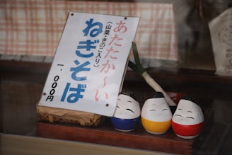 A Japanese sign featuring bold, blue and red text is displayed, leaning against a basket. In front of it are three small, painted statues with cheerful facial expressions, each a different color: blue, yellow, and red. A leek is positioned upright behind the statues, serving as a decorative element.