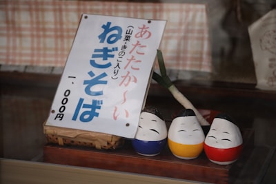 A Japanese sign featuring bold, blue and red text is displayed, leaning against a basket. In front of it are three small, painted statues with cheerful facial expressions, each a different color: blue, yellow, and red. A leek is positioned upright behind the statues, serving as a decorative element.