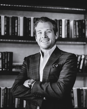 A monochrome photograph of a smiling person in a plaid suit with arms crossed, standing in front of shelves filled with books.