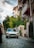 A vintage white car is parked on a cobblestone street flanked by old, charming buildings with balconies. Lush greenery surrounds the area, adding a serene and nostalgic atmosphere to the scene under a partly cloudy sky.