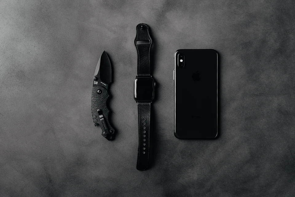 The image presents a scene on a gray concrete surface, with three objects arranged in a triangular formation. On the left side is a black knife with a curved blade and a handle. In the center is an Apple watch with a silver band and a black face. On the right side is a black iPhone with a white back cover.